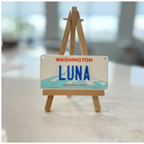 Personalized 4x2 inch mini Washington state license plate displayed on a wooden tripod stand for product presentation State Plate for Kids, Car Toy, Wagon, Bike, Stroller – Thick Plastic Name Sign