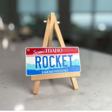 Miniature Idaho license plate with 'Rocket' on a wooden stand against a blurred background. Personalized Mini License Plate Tag – Custom 4x2” State Plate for Kids, Car Toy, Wagon, Bike, Stroller – Thick Plastic Name Sign.