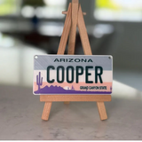 Arizona license plate with 'COOPER' on a wooden stand against a blurred background. Personalized Mini License Plate Tag – Custom 4x2” State Plate for Kids, Car Toy, Wagon, Bike, Stroller – Thick Plastic Name Sign.